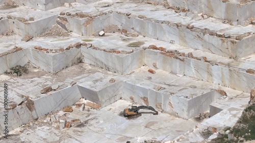 SICILY, ITALY, 24 JAN 2026: AERIAL, CLOSE UP: Excavator sits on a bright marble bench surrounded by huge cut walls and scattered stone blocks. Modern quarry operations and marble industry in Sicily.