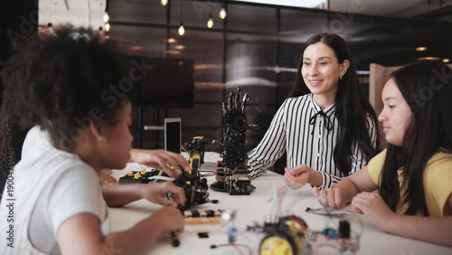 White female teacher advises and instructs primary girls group students, Robotic arms design at learning table in science classroom, discusses experiment knowledge for children of elementary school.