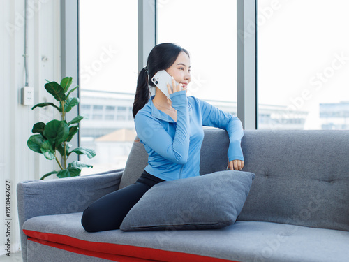 Asian young beautiful happy woman fit sporty active healthy in sportswear sitting on cozy sofa using calling mobile smartphone after workout exercise training in living room at home wellness lifestyle