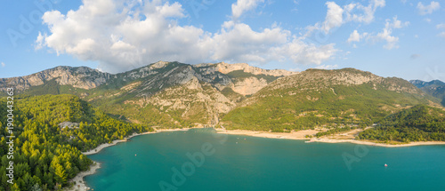 Aerial view of Lac de Sainte-Croix with vivid turquoise water, sandy shoreline, and rugged limestone mountains under a bright sky with scattered clouds.