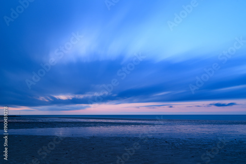 Konstfotografi Long exposure captures streaked blue clouds and tranquil water at low tide along the English Channel, with soft pastel light fading on the horizon and wet sand reflecting the sky