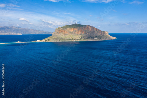 A striking rocky outcrop rises from the brilliant blue Mediterranean waters near Monemvasia, Greece, under a bright sky with scattered clouds. Sunlight enhances the rugged textures and natural colors