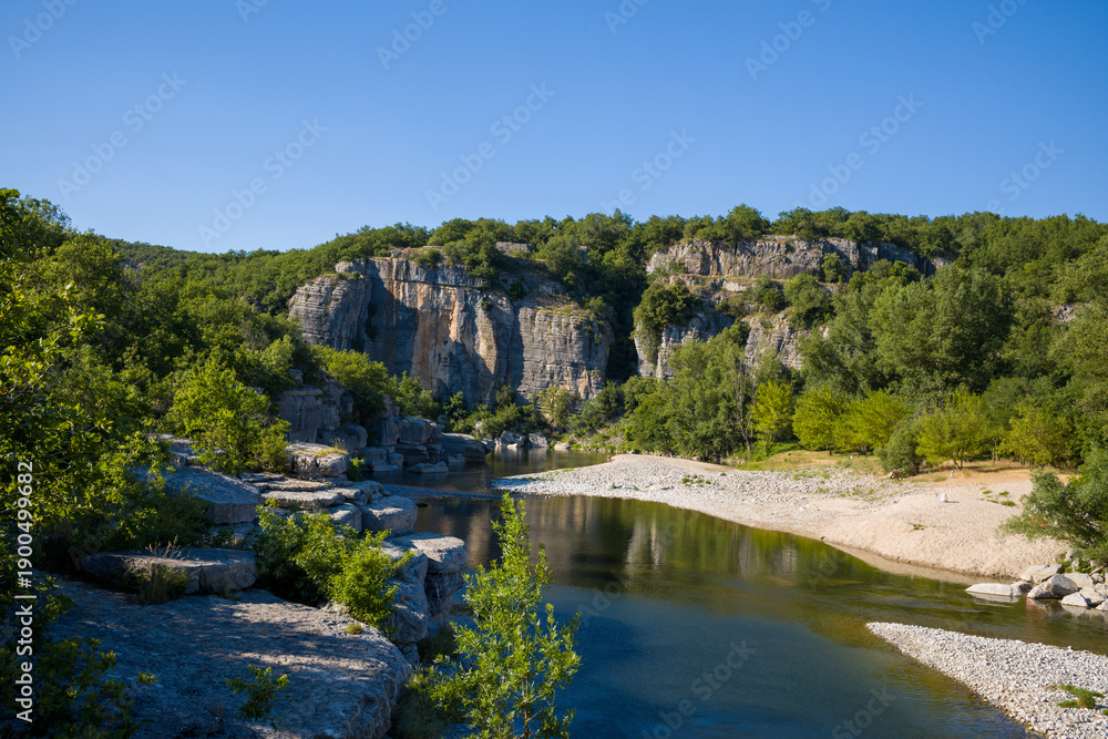 Fototapeta premium Clear river winding through dramatic limestone gorges, with rocky outcrops and lush green vegetation under a vivid blue sky in the Gorges de l'Ardeche.