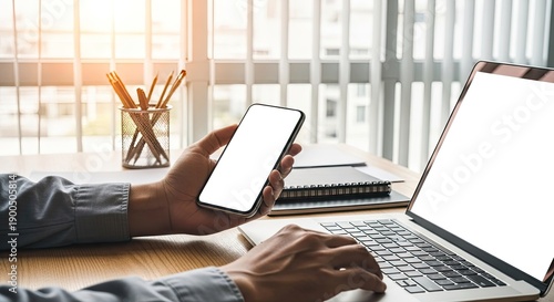 Person using a smartphone and laptop at a wooden desk in an office with bright sunlight streaming