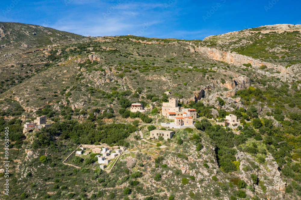 Fototapeta premium Aerial view of traditional stone buildings nestled among rocky slopes and green vegetation on a hillside in Mani, Greece, under a clear blue sky.