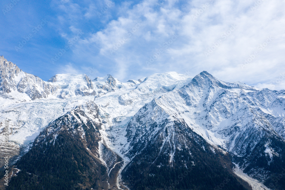 Obraz premium Expansive view of the Mont Blanc massif with rugged snow-covered summits and forested lower slopes, illuminated by soft daylight beneath a partly cloudy sky.
