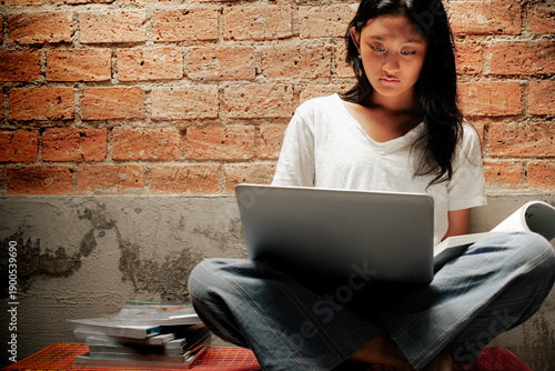 Asian teenage girl sitting cross-legged reading a book with a laptop on her lap against a red brick wall background, studying online at home.