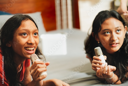 Two girls lying on a bed singing into microphones, smiling and laughing together during a fun karaoke moment at home.