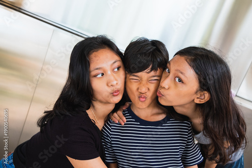 Two girls making kissy faces at an annoyed boy making a funny scrunched expression between them, playful sibling teasing moment indoors.