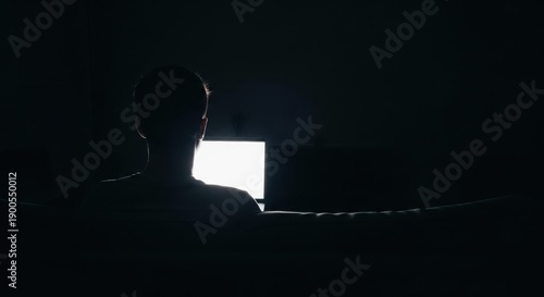 Man standing in front of a computer screen with a bright light in a dark room from behind