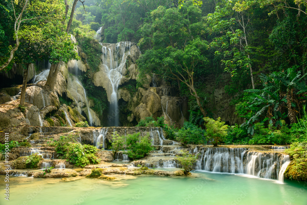 Naklejka premium Layered limestone cascades flow into vibrant turquoise pools surrounded by dense tropical forest at Kuang Si Waterfalls in northern Laos. Sunlight highlights the lush greenery and natural textures