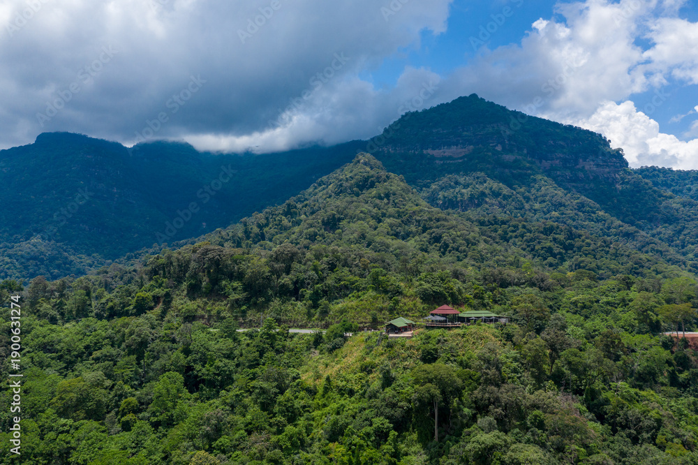 Naklejka premium A small lodge with red and green roofs sits nestled in dense tropical forest on a verdant hillside, with dramatic mountain peaks and low clouds in the background. The scene captures the rich greenery