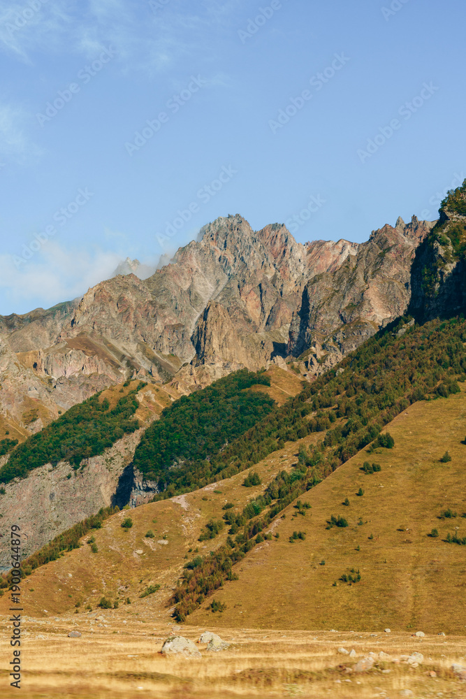 Fototapeta premium Rocky mountains rise above grassy fields in the daytime sunshine
