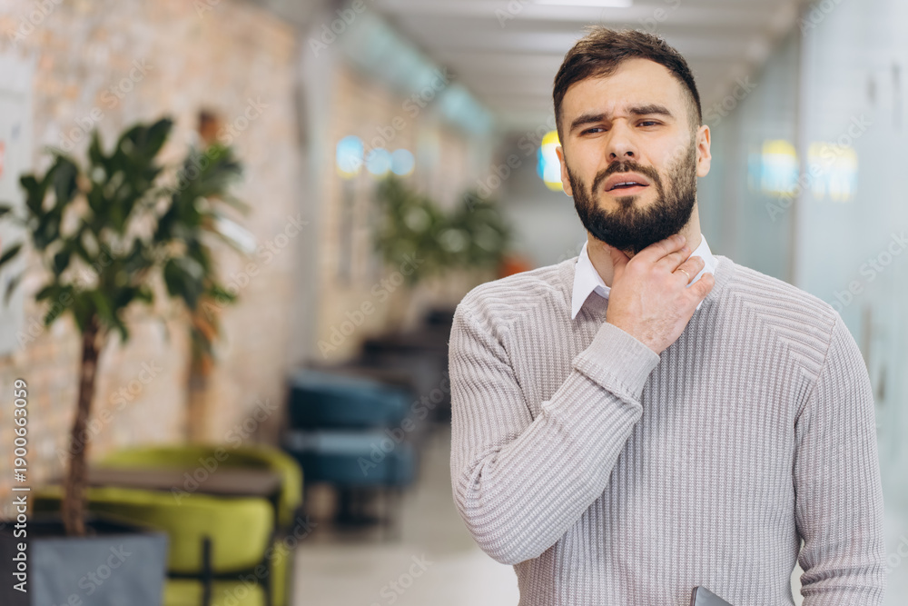 Naklejka premium Man suffering from a sore throat and discomfort, touching his neck with a pained expression in an office environment