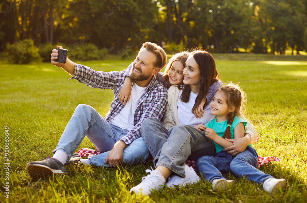 Fototapeta premium Smiling parents with two children sitting on grass, hugging and taking selfie together in sunny park. Happy family of four enjoying quality time outdoors, capturing cheerful moment with camera.