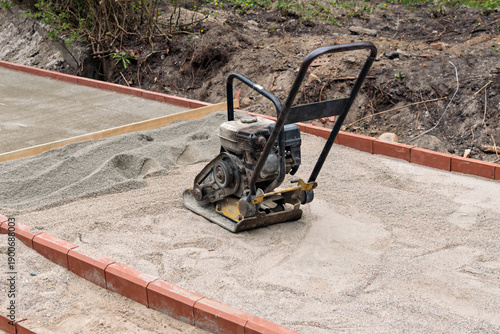 Canvas Print Plate compactor rests on sand base inside curb as paving walkway is prepared during renovation at landscaping construction site