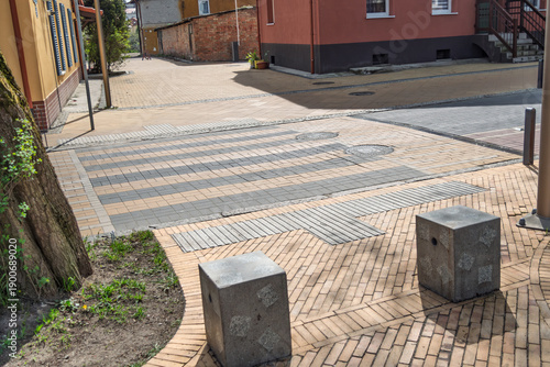 Quadro em tela Pedestrian crossing in quiet courtyard features patterned brick paving, tactile guidance strips, and concrete bollards in sunlight