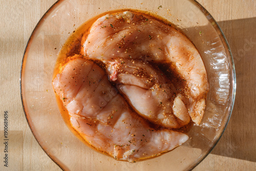 Raw chicken breasts marinate in spicy herb oil inside glass bowl on wooden board in warm sunlight, top view for home cooking prep.