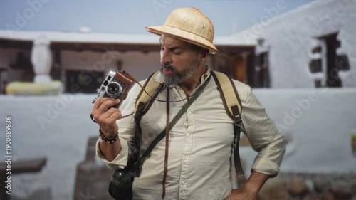 Man holding vintage camera, finger to ear and hands visible at building entrance wearing pith helmet and backpack; confusion.