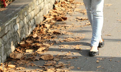A person walking along a leaf-strewn pathway in light jeans and black flats, beside a stone wall on a crisp autumn day