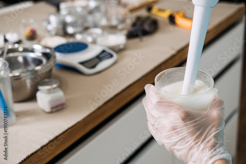 Person using immersion blender to mix liquid base in glass jar on digital scale. cosmetic emulsifying, professional cream blending, skincare laboratory tools, making lotion process.