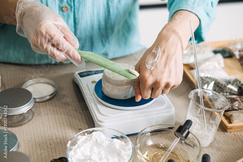 Fotografia Person in gloves applies cream from spatula to hand while measuring jar on digital scale