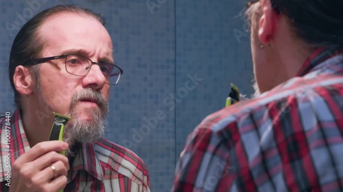 Close-up of a middle-aged man in the bathroom, meticulously trimming his beard away with an electric shaver to adopt a new look and style by keeping only his mustache