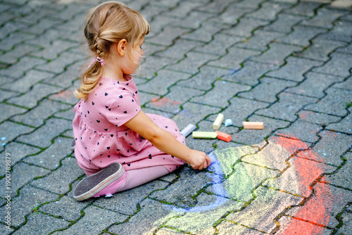 Little preschool girl painting rainbow with colorful chalks on ground on backyard. Positive happy toddler child drawing and creating pictures. Creative outdoors activity in summer.