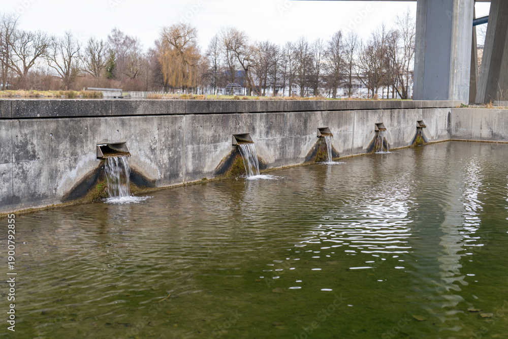 Naklejka premium Flowing water from several rectangular spouts enters a green water basin near a bridge during winter day
