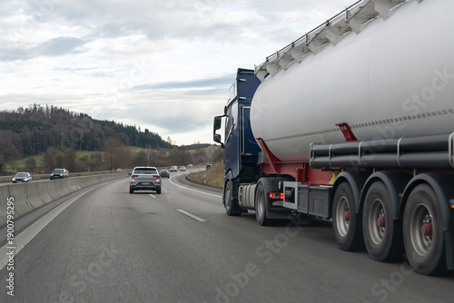 Large blue semi-truck with a white tanker trailer driving on a multi-lane highway with other cars under an overcast sky