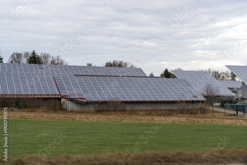 Large solar panels cover multiple barn roofs on a cloudy day, set against a green field and distant trees in the countryside