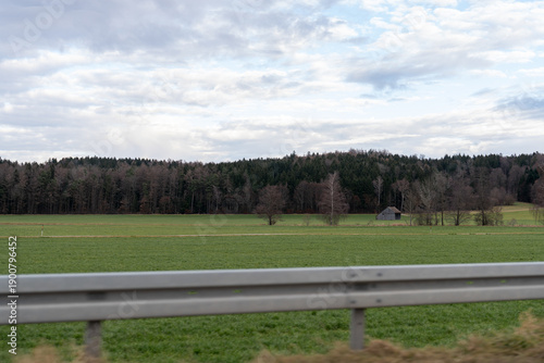 Roadside view of a vast green field, dark forest line, and a remote wooden shed under cloudy skies