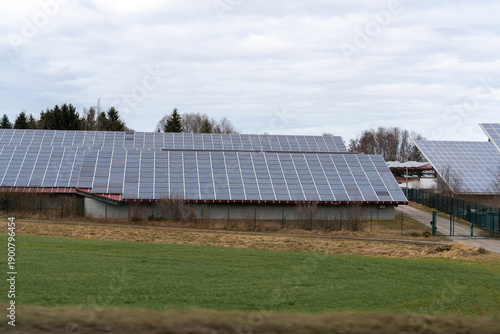 Solar panels covering multiple large barn roofs near a green field and evergreen trees on an overcast day