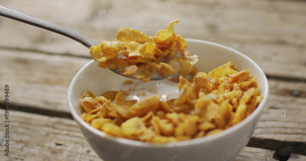 Fototapeta premium Scooping metal spoon pulling golden corn flakes from white china bowl on wood planks, milk clinging