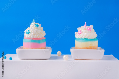 Layered cheesecakes with whipped cream decorated with unicorn themed sugar sprinkles against the blue background. Two desserts for kids party or gender reveal party