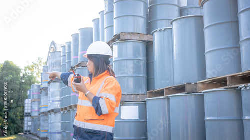 A worker in safety gear is using a walkie-talkie while pointing towards the stacked barrels. The scene takes place in a warehouse with good lighting