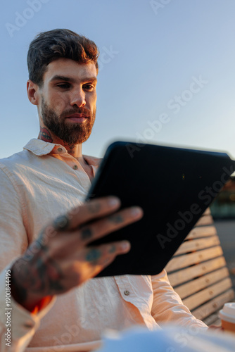 Man with beard using tablet on a bench outdoors at sunset, conveys focus and modern lifestyle
