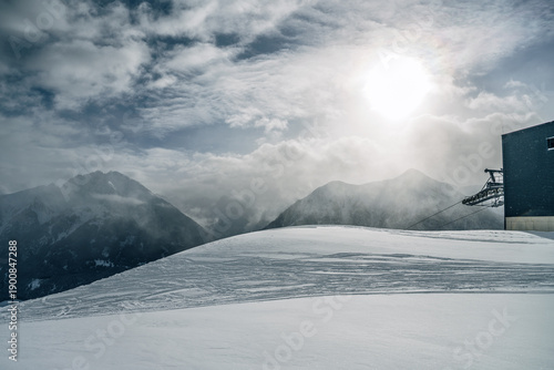Fotografie Sun breaking through clouds at a snowy mountain summit station, dramatic winter light, alpine peak, fresh snow, atmospheric and cold winter scene