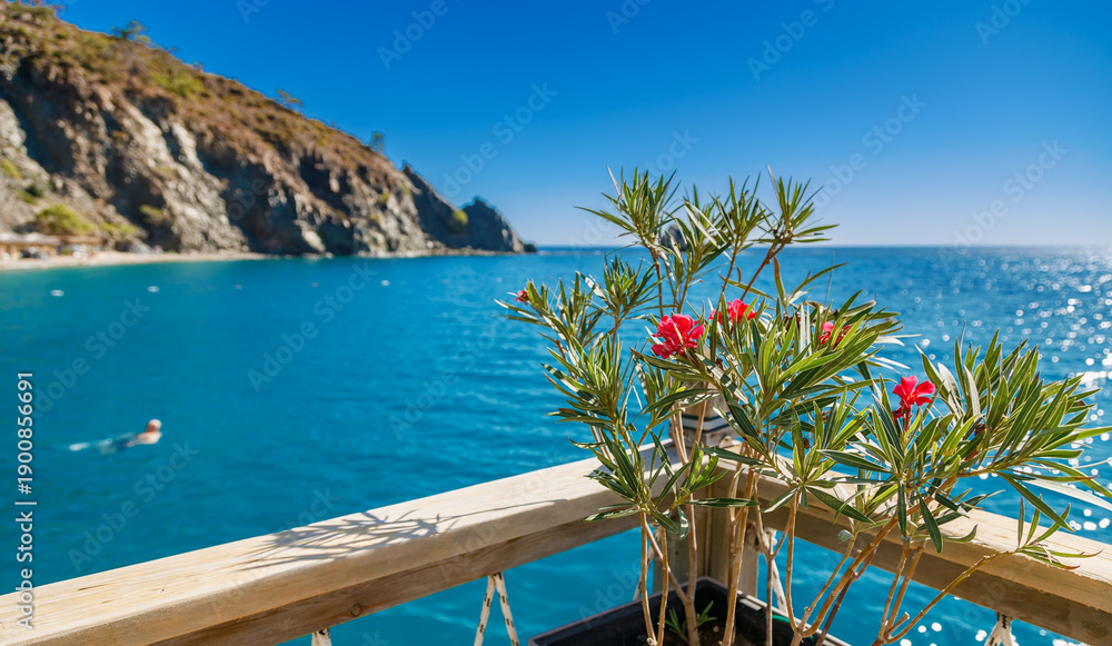 Fototapeta premium Beautiful mediterranean sea view from a wooden balcony with blooming pink oleander flowers and rocky cliffs under a clear blue sky for luxury travel and summer vacation concepts
