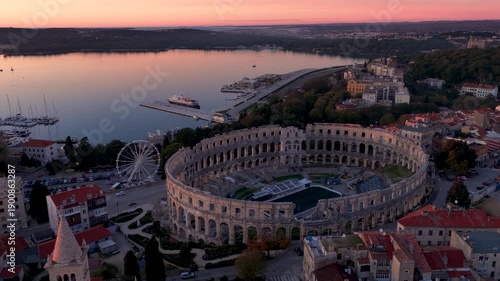 Aerial view of the ancient Pula Arena, a grand amphitheater, juxtaposed with the serene harbor under a soft, pastel sky, Pula, Istria County, Croatia.