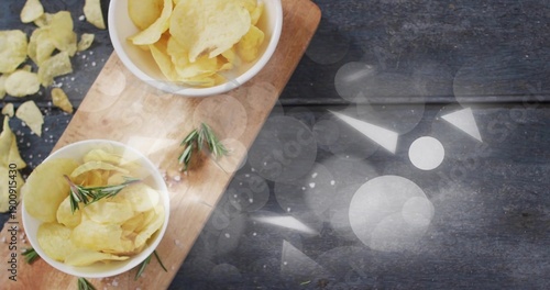 Showing two white bowls holding potato chips on cutting board at kitchen table with rosemary, salt