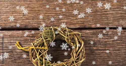 Fototapeta Displaying grapevine wreath with moss and yellow twine on weathered planks, scattering snowflakes