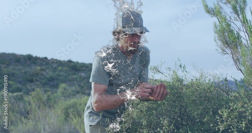 Examining man bending holding plant in scrubby hills, wearing cap and T-shirt with starry overlay