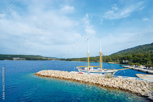 Obraz na plátně A sailboat docked at a rocky pier on a clear blue sea, surrounded by lush green hills under a partly cloudy sky
