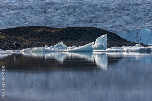 Icebergs drift in quiet harmony across Jökulsárlón glacier lake as soft evening light settles over Southern Iceland’s rugged dark ridges and the distant glacier shimmering behind them