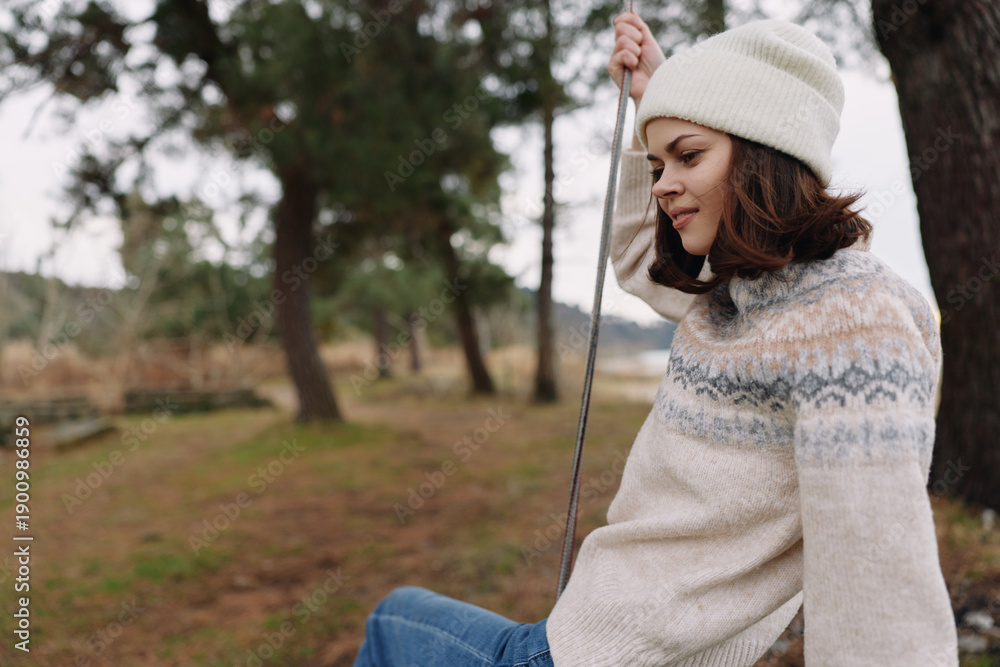 Fototapeta premium Woman sitting on a swing in a park, wearing a knitted sweater and beanie, relaxing outdoors. Serene casual scene with trees and soft light, perfect for lifestyle, nature, and seasonal moments.