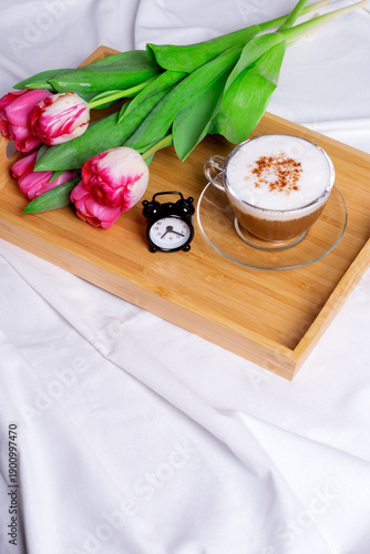 Cappuccino with cinnamon, alarm clock and pink tulips on the wooden tray in bed. Waking up and small morning rituals