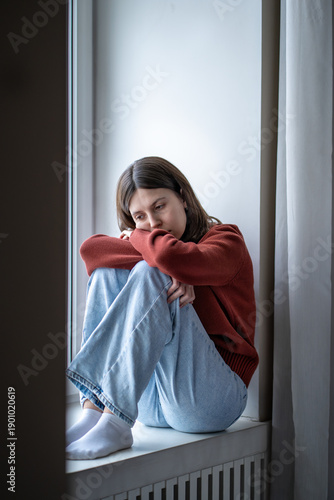 Konstfotografi Depressed lonely young woman sitting on windowsill sadly looking feeling anxiety