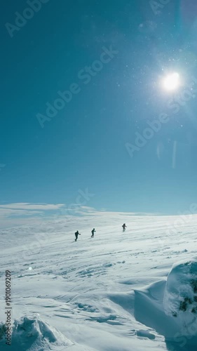 Three people are skiing on a snow-covered mountain. The sky is clear and blue, and the sun is shining brightly. Scene is peaceful and serene, as the skiers enjoy the beautiful winter landscape