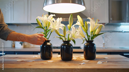 Hands of women work together to arrange flowers in black vases on a wooden countertop in a bright kitchen. Petals are scattered around as they focus on the task in the warm light of daytime